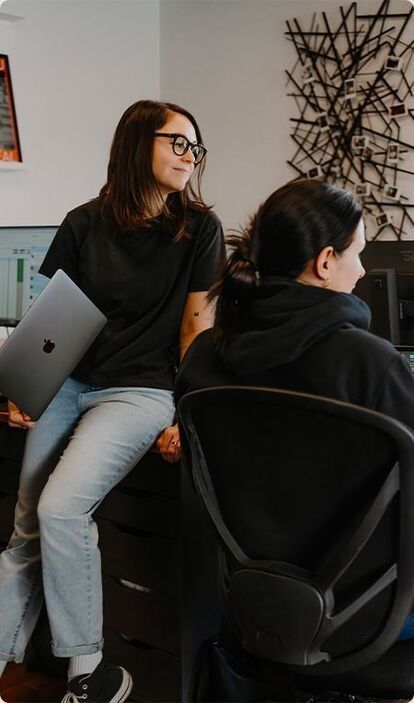 Deux employées, assises côte à côte dans un bureau à Mirabel, sur la Rive-Nord de Montréal, se réunissent autour d'un ordinateur. Elles discutent d'un mandat créatif et finalisent les détails d'un projet.