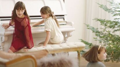 Two young girls are sitting near a white piano. A Christmas tree stands in the house. The walls are white, and a third little girl is sitting on the floor.