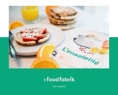 A plate of breakfast bread topped with strawberries, shredded coconut and syrup, accompanied by a glass of orange juice and orange slices. Next to it, a “L'ensoleillé” food packaging with the words “la food fabrik” and “bon appétit”.