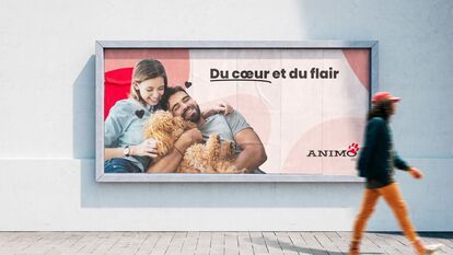 Billboard mounted on the white wall of a pale gray brick alley. The billboard features two people seated and intertwined, with a beige curly-haired dog in their arms. The image features the ANIMO logo and the text “Du cœur et du flair” and “été”.