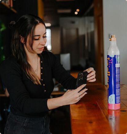 A social media manager, cell phone in hand, stands in front of a client's product. She takes photos and videos to create engaging content for the agency's client's social networks. 