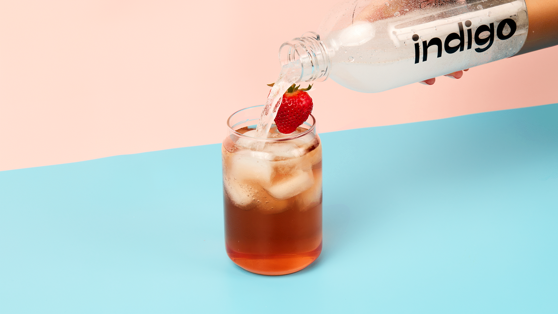 Une main verse de l'eau pétillante d'une bouteille en plastique transparente dans un verre rempli de glaçons et de thé glacé d'une teinte rosé. Une fraise est posée sur le bord du verre. Le fond est divisé en deux couleurs, rose et bleu.
