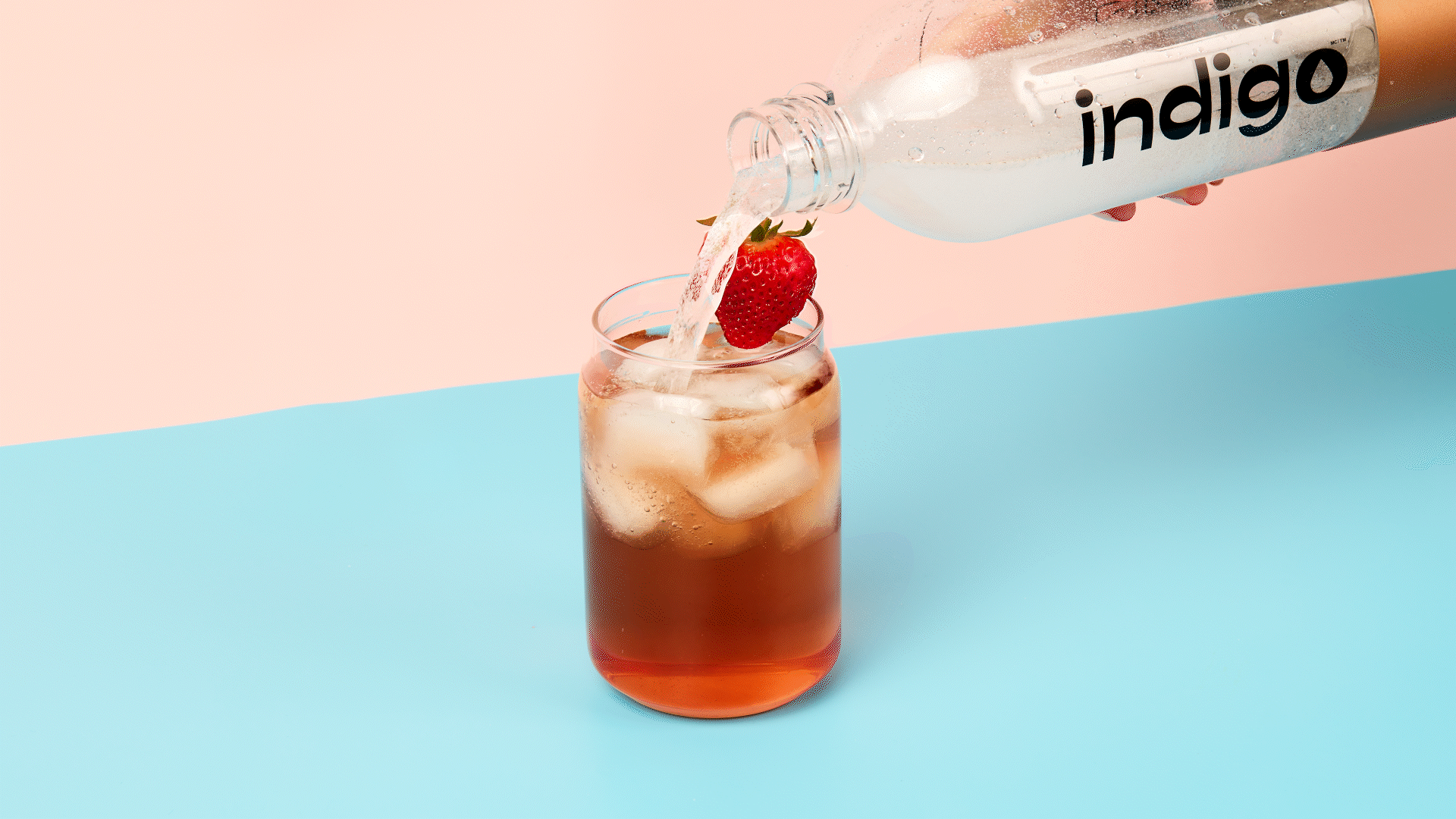 A hand pours sparkling water from a clear plastic bottle into a glass filled with ice cubes and iced tea of a rosy hue. A strawberry sits on the rim of the glass.