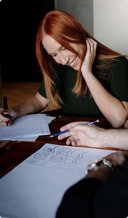 Une jeune femme souriante, assise à un bureau moderne, collabore activement avec son équipe sur un projet. Des feuilles blanches avec des maquettes de design sont étalées sur la table, témoignant du processus créatif en cours.