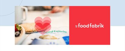 A “La Food Fabrik” product package is shown in close-up on a table, next to a plate containing a roll, strawberries and blueberries. The “La Food Fabrik” logo appears in red on a plain background, with three pink hearts superimposed in the background.